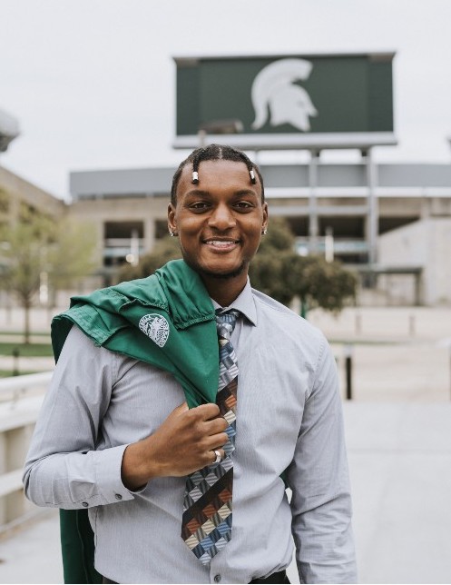 A young man with a blue dress shirt and tie holding a green graduation gown in front of a green MSU Spartan sign
