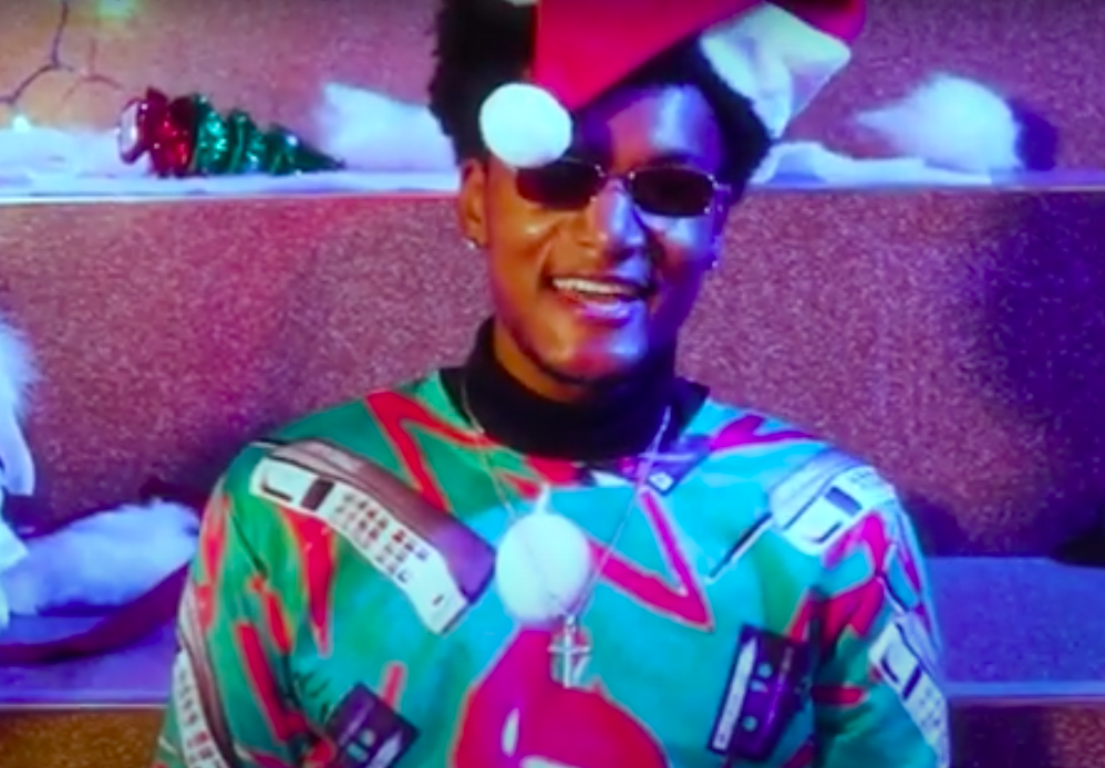 a screenshot of a video with a young man in a Santa hat and a holiday shirt wearing sunglasses