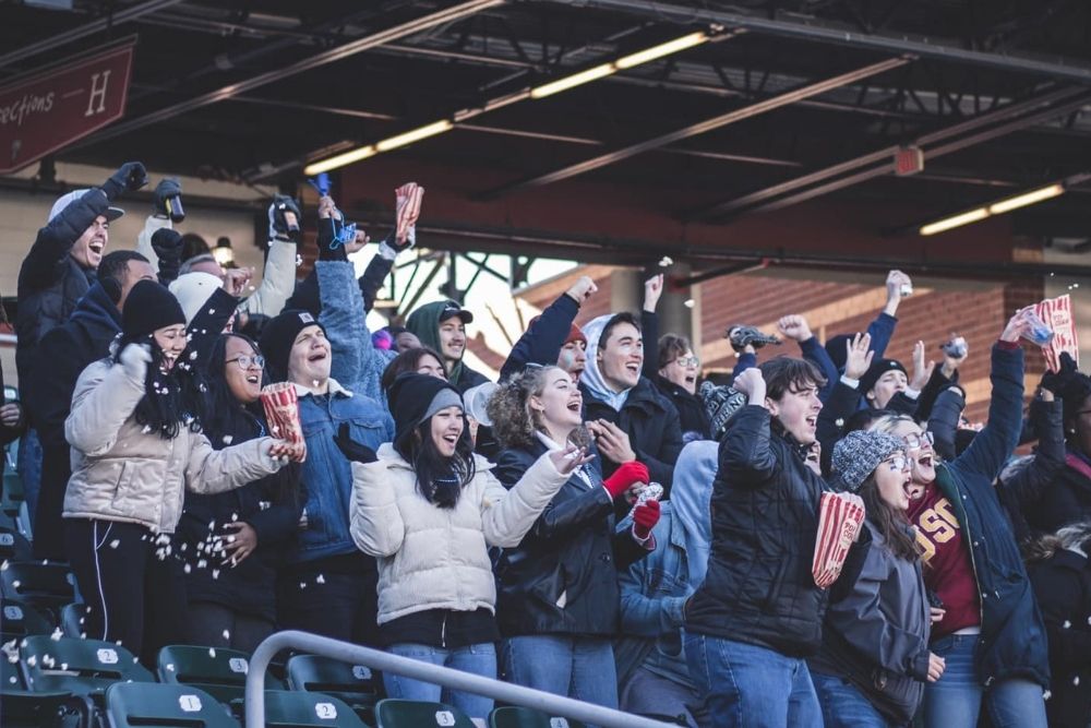 A crowd of young people with popcorn cheer in the stands of a baseball stadium.