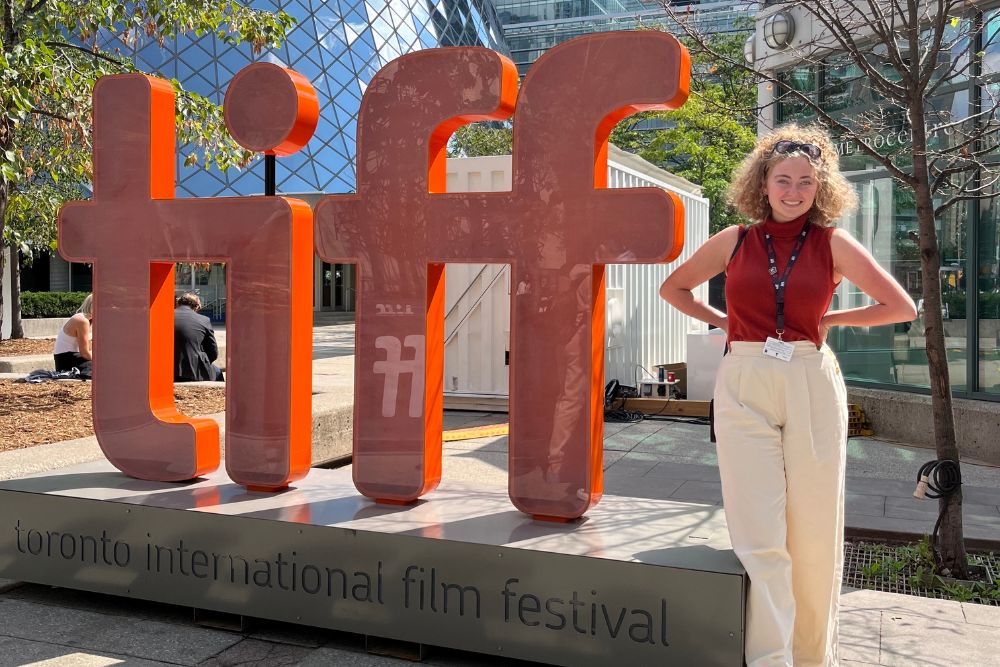 Young woman stands outdoors beside a large, orange decoration sign for the Toronto International Film Festival.
