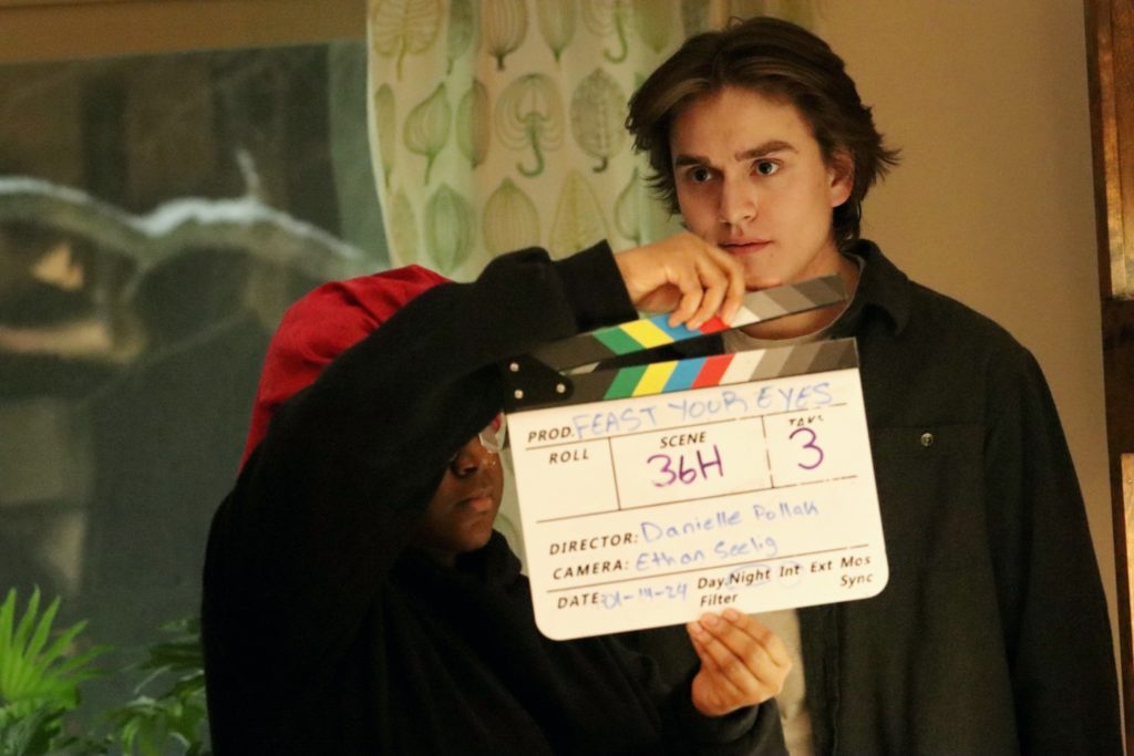 A student stands in front of an actor holding a clapperboard on the set of Feast Your Eyes.